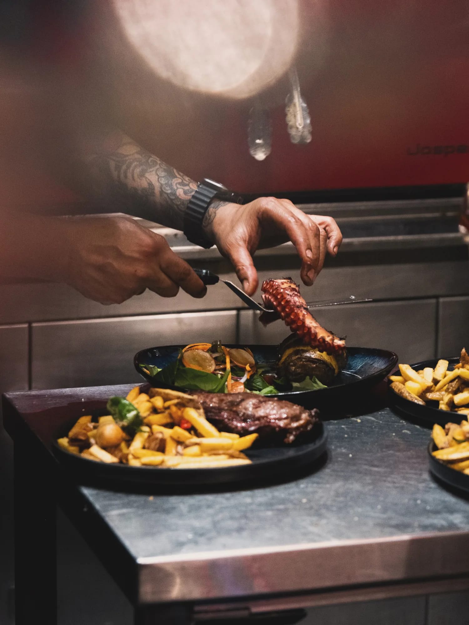 A chef plating grilled octopus in the kitchen, the Josper behind him and plates of skirt steak with fries