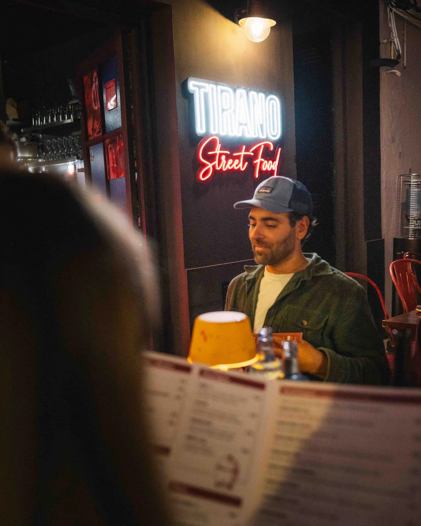 A customer at the Tirano bar under the neon sign, menu open in front of him and a beer on the table