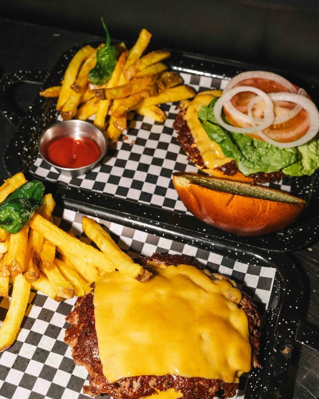 Two smash burgers with melted cheese served on trays with fries and padrón peppers