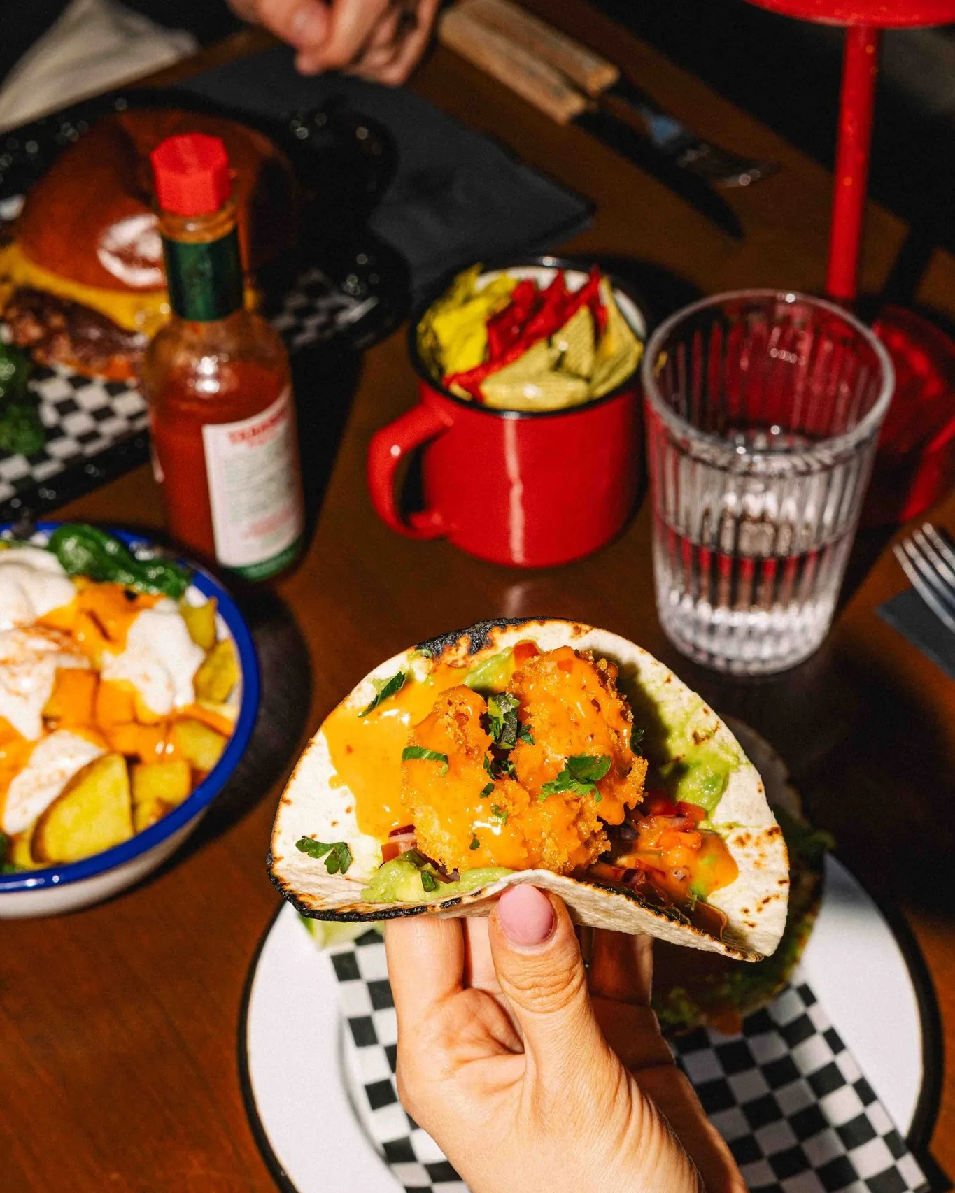 A hand holding a crispy cauliflower taco with guacamole, over a table with hot sauce and potatoes